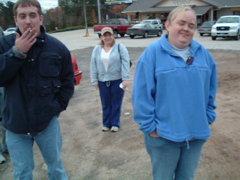 Three people standing in a dirt parking lot with vehicles in the background
