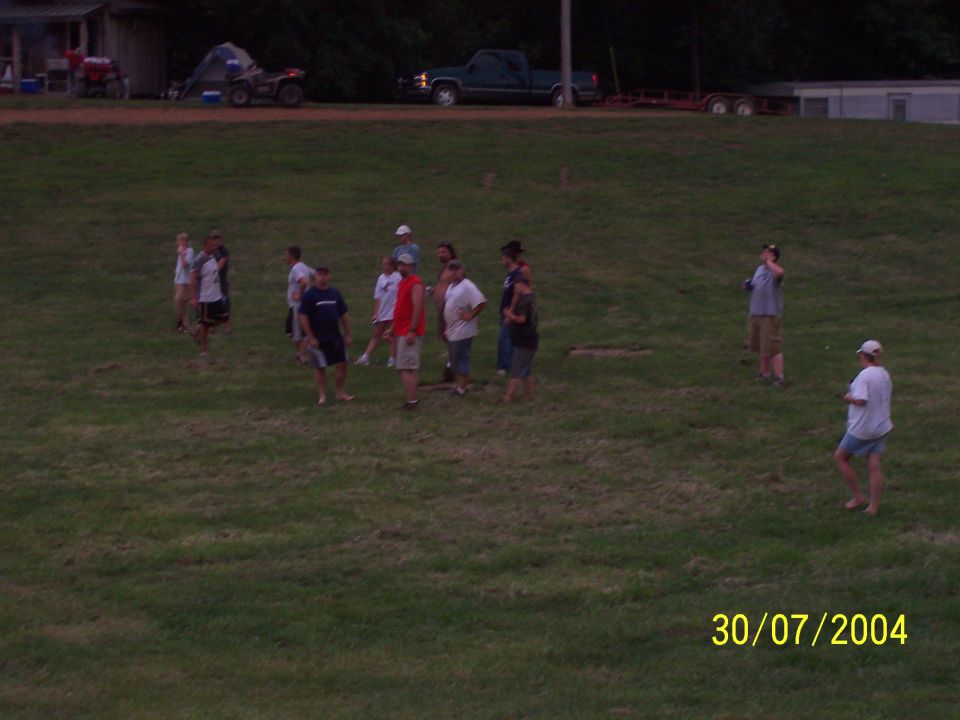 Group of people gathered in a grassy field with a building and vehicles in the background