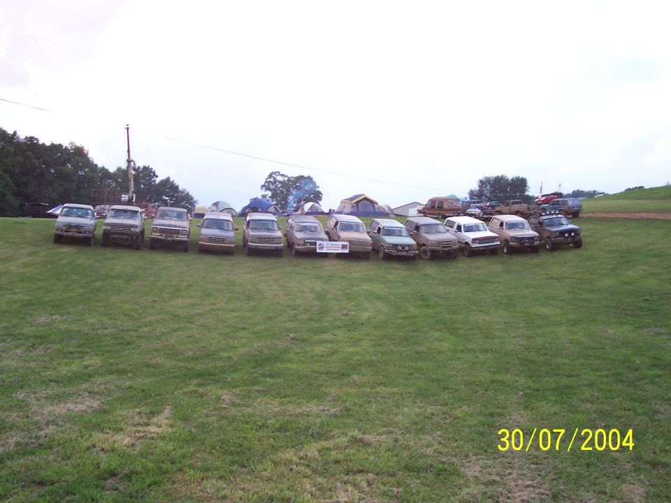 Line of older trucks and SUVs parked on grass, tents in background
