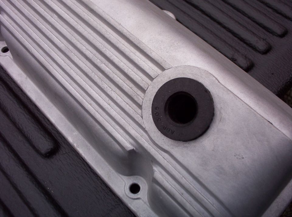 Aluminum engine valve cover with ribbed texture and a black grommet. Text 'ADP 303' is visible