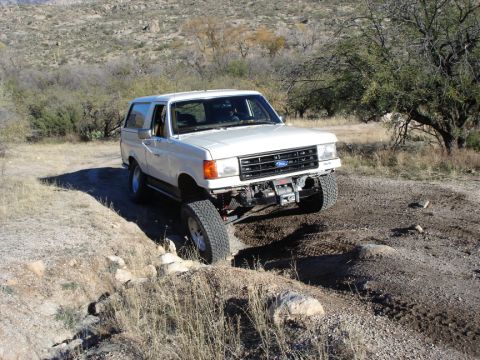 1987 Ford Bronco - A white 1987 Ford Bronco navigates a challenging off-road trail with one wheel lifted.