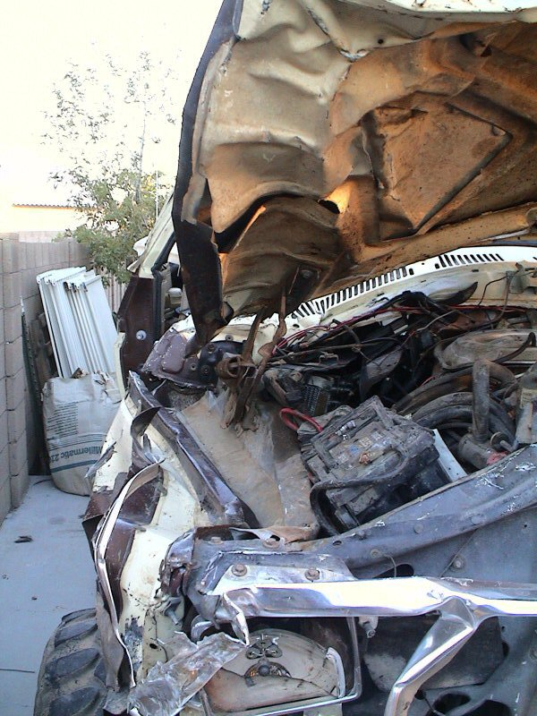 Crushed front end of a white truck with folded rusty hood, torn wiring and bent chrome bumper