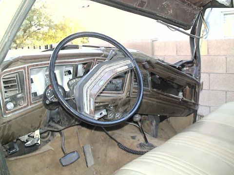 1979 Ford Bronco - Interior of a stripped vintage Ford truck shot from the passenger side, showing a black four-spoke steering wheel, woodgrain-trimmed dash w…