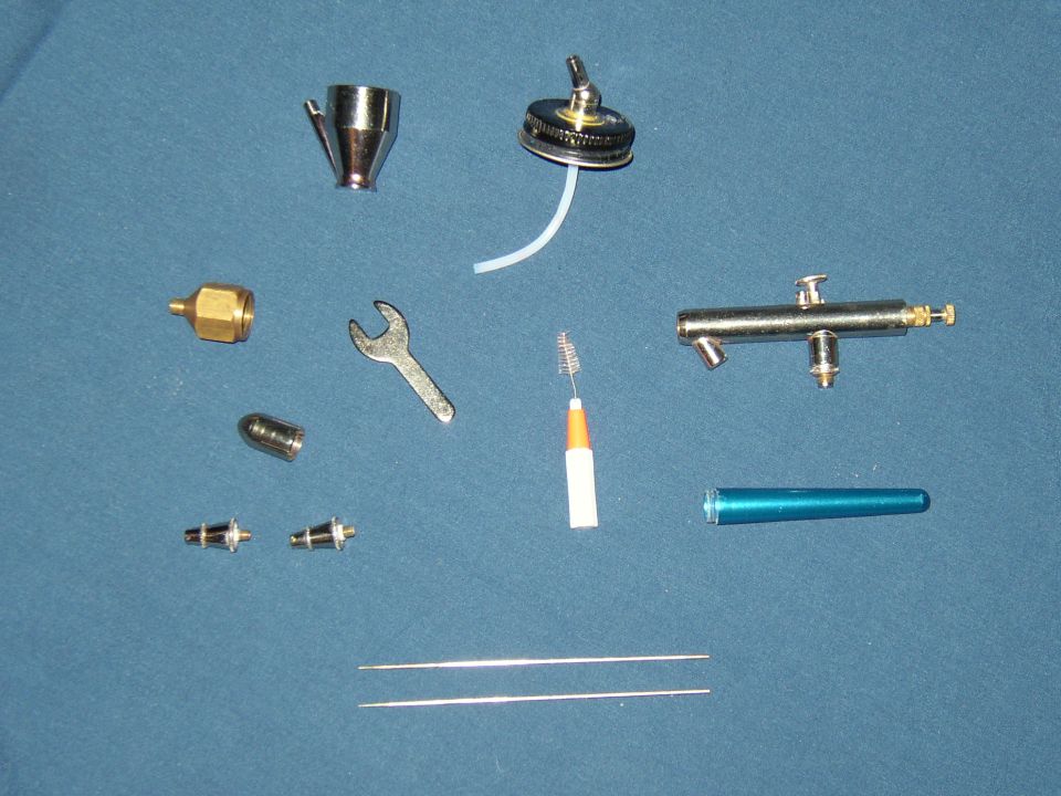 Airbrush parts laid out on blue surface, including nozzles, a wrench, cleaning brushes, and a metal tube