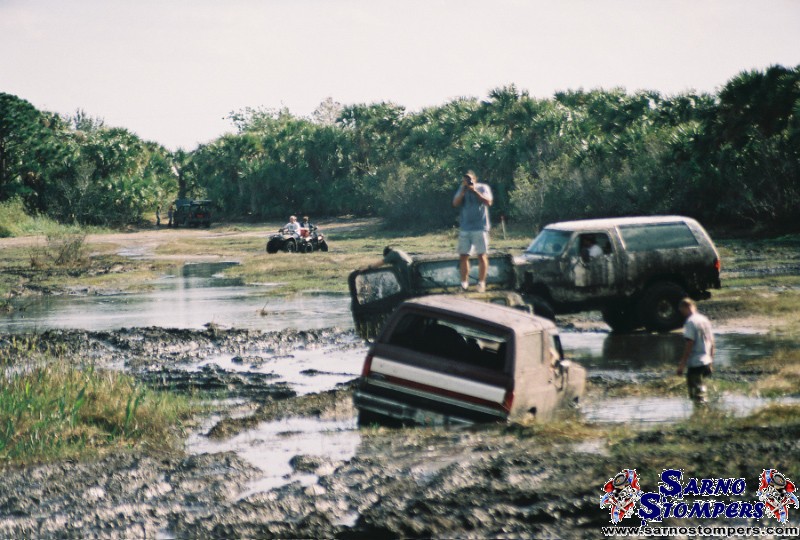 Several off-road vehicles and people at a muddy bog, one truck sunk to its windows in the water