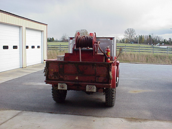 Rear view of a red fire truck with a hose reel and extinguisher mounted in the bed, parked in front of a garage