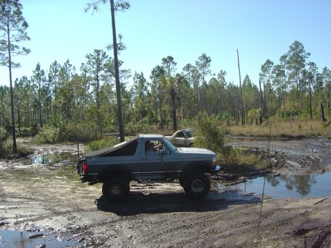1993 Ford Bronco - Ford Bronco navigates a wet, muddy trail surrounded by trees under a clear blue sky.