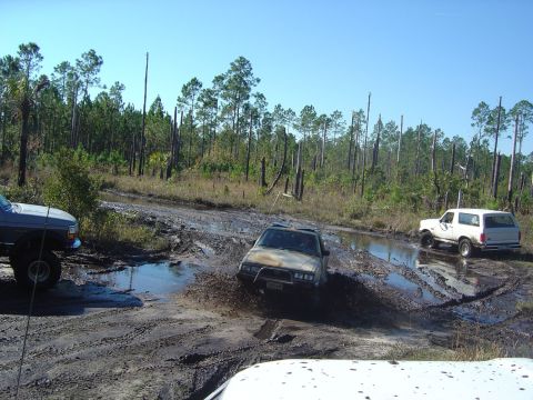 1993 Ford Bronco - Off-road vehicles navigate a challenging muddy terrain with trees in the background under a clear blue sky.