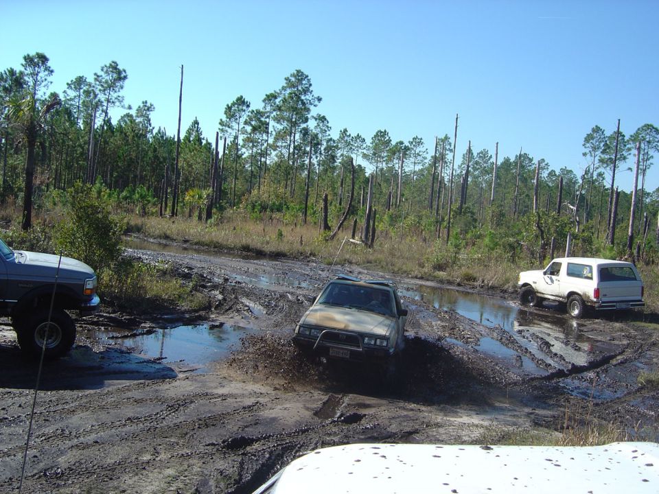 Several SUVs driving through a muddy, wooded area. One SUV is splashing through a deep puddle