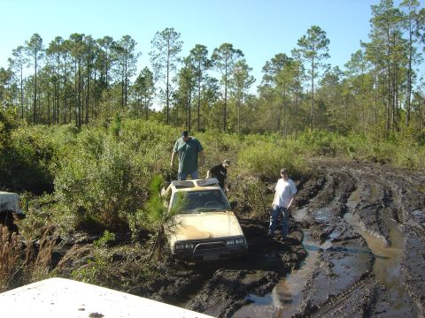 1993 Ford Bronco - Three people stand around a yellow off-road vehicle that is deeply stuck in a muddy track.