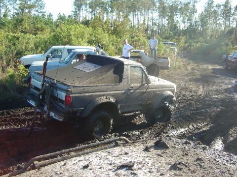 1993 Ford Bronco - Multiple trucks, including a mud-caked 1993 Ford Bronco, are stuck in a muddy off-road environment.