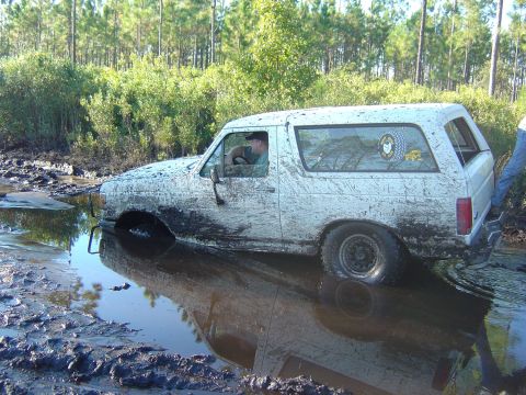1993 Ford Bronco - Muddy 1993 Ford Bronco is stuck in a water-filled ditch with trees in the background.