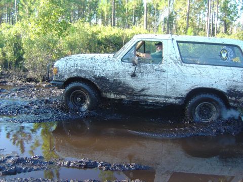 1993 Ford Bronco - A white Ford Bronco navigates a challenging muddy terrain, with trees in the background.