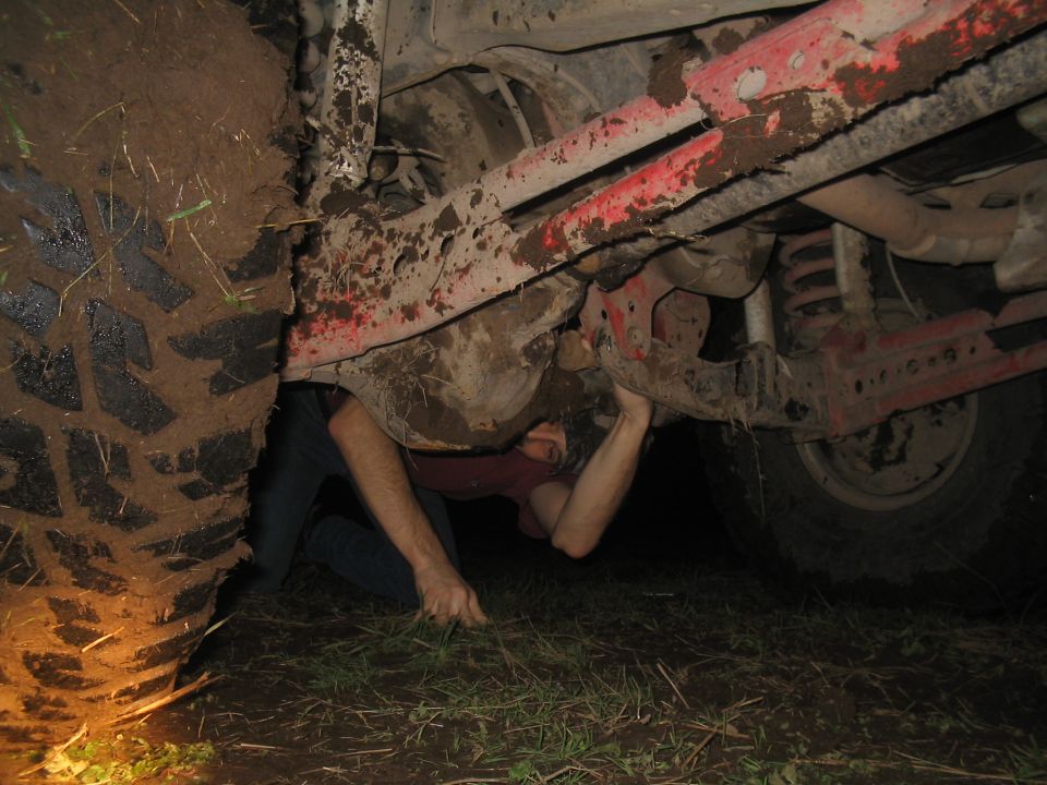 Person working on the muddy undercarriage of a vehicle, near a large tire