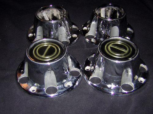 Four chrome hubcaps with gold centers and visible lug nut holes, on a dark surface