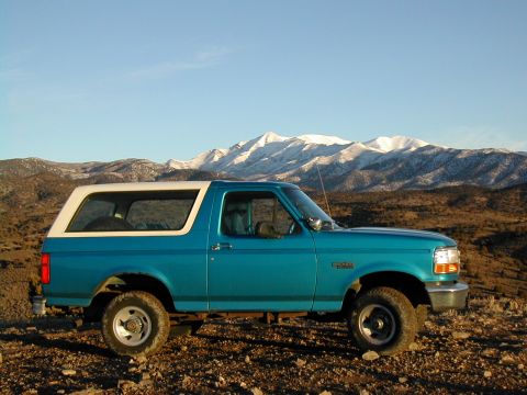 1994 Ford Bronco - A teal Ford Bronco with a white top is parked on a rocky, arid landscape with snow-capped mountains under a clear blue sky.