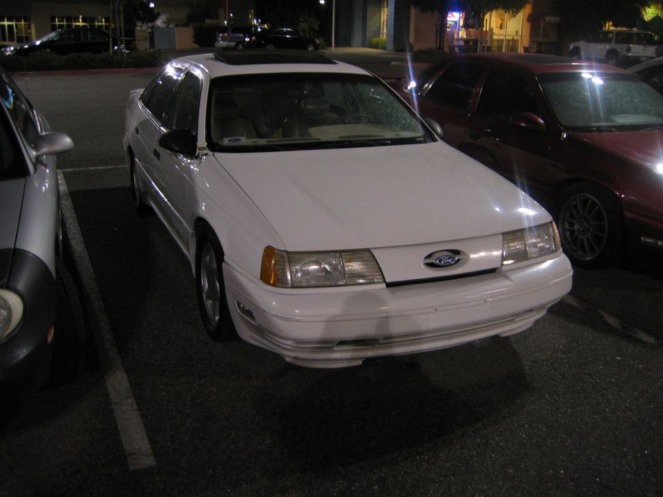 White sedan parked at night. It has a sunroof and a Ford badge on the grille. A red car is parked next to it