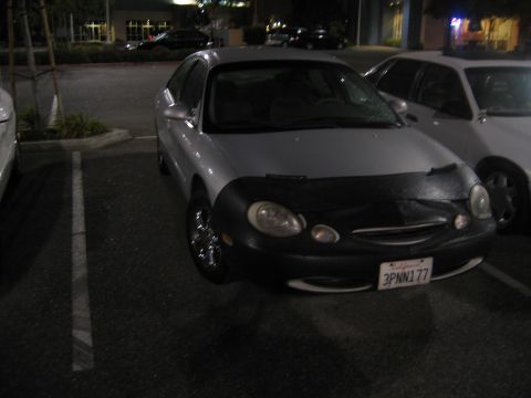 2002 Ford Taurus - A silver sedan with a black car bra is parked in a lot at night, with another vehicle visible to its right.