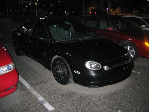 2002 Ford Taurus - A black sedan is parked in a lot at night, with other vehicles nearby. The car has custom wheels and stickers.