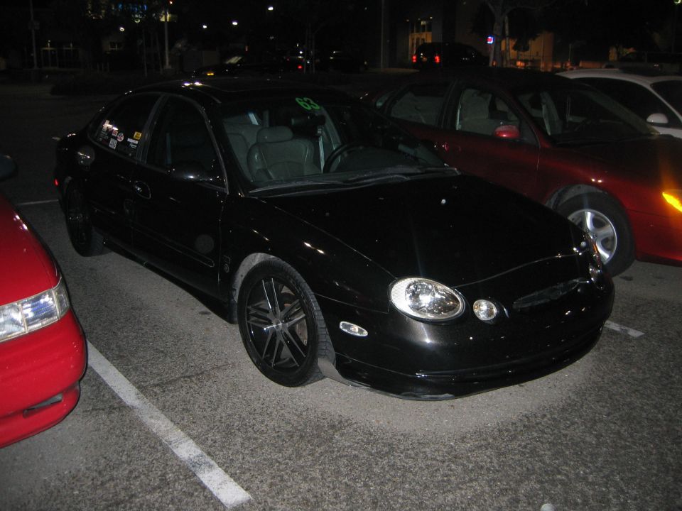 Black sedan parked at night, red car to the left, red car to the right. Has stickers on the driver's side door