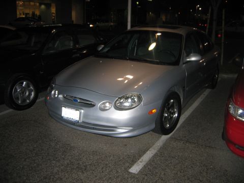 2002 Ford Taurus - A silver 2002 Ford Taurus is parked between a black car and a red car at night.