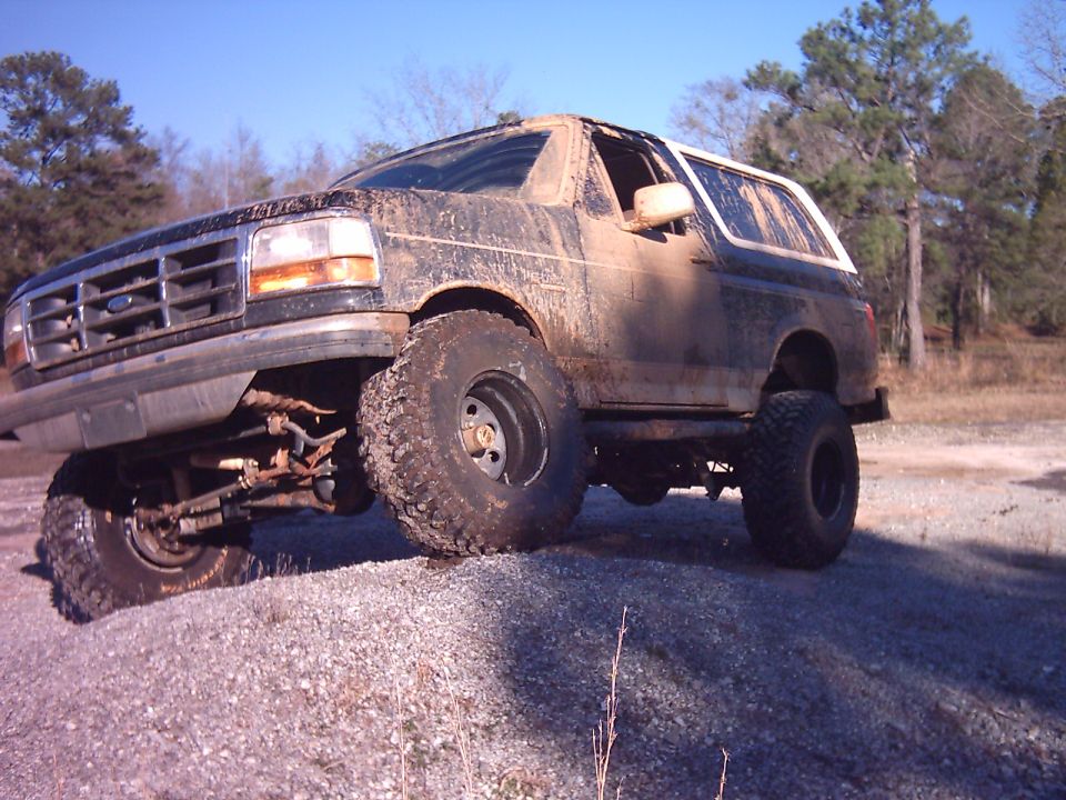 Muddy lifted Ford Bronco on large mud tires parked on gravel, front passenger side raised, trees behind