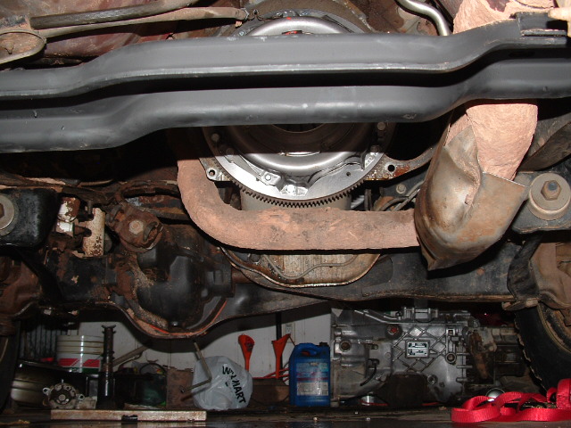 Underside view of vehicle showing exposed flywheel/ring gear and rusty drivetrain components in a shop