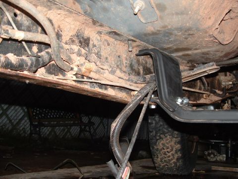 1990 Ford Bronco - Underside view showing a rusty vehicle frame rail, a black plastic guard or tank, wiring loom and mud tire in a garage.