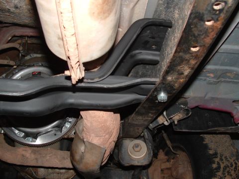 1990 Ford Bronco - Underbody view of a vehicle showing a black-painted crossmember or sway bar, a hex-bolt frame mount, and various rusty chassis components w…