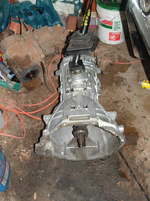 1990 Ford Bronco - A manual transmission removed from a vehicle, resting upright on a concrete garage floor with the shifter and rubber boot still attached an…