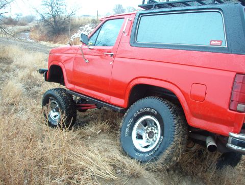1985 Ford Bronco - This lifted 1985 Ford Bronco is shown in a rural outdoor setting with dry grass and a stream in the background.
