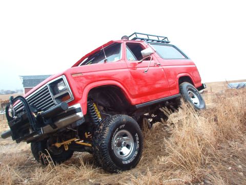 1985 Ford Bronco - This lifted SUV is navigating a grassy, uneven terrain with a clear sky overhead.