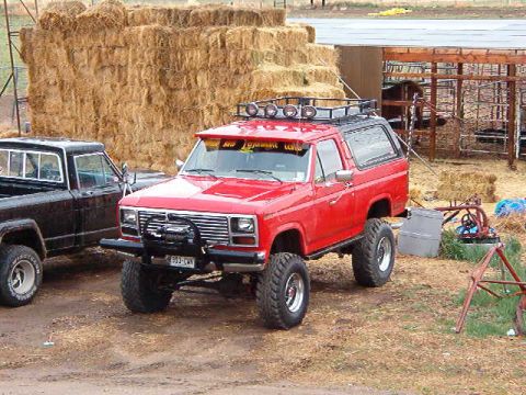 1985 Ford Bronco - Red Ford Bronco parked next to a black pickup truck on a dirt lot with hay bales and a wooden structure.