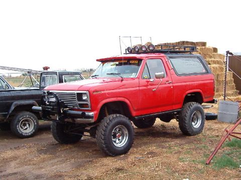 1985 Ford Bronco - A red SUV with off-road tires and a roof rack is parked outdoors next to another vehicle.
