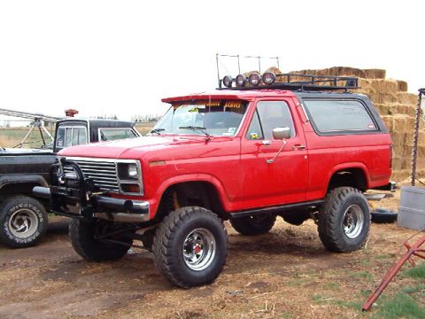 1985 Ford Bronco - A red Ford Bronco, modified with a lift kit and extra lights, sits beside a black truck.