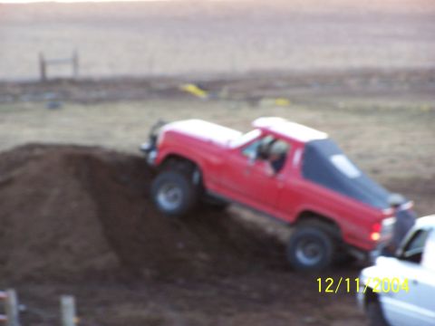 1985 Ford Bronco - A red SUV with a black soft top navigates a dirt hill, with another vehicle partially visible.