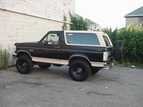 1993 Ford Bronco - Driver-side profile of a lifted black full-size Ford Bronco with a tan removable hardtop and matching tan lower body stripe, parked on crac…