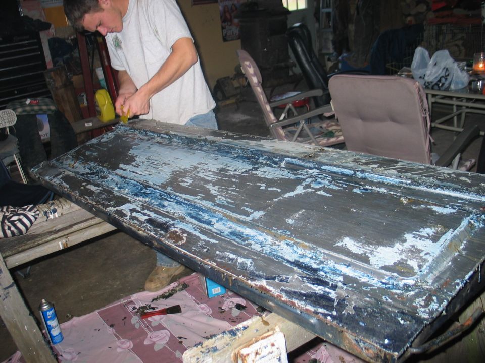 Man stripping paint from a long metal body panel on sawhorses in a garage