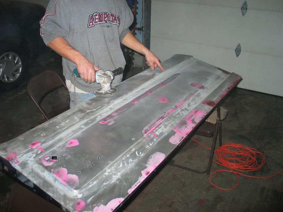 Person sanding a bare-metal tailgate with pink body filler spots on folding chairs in a garage