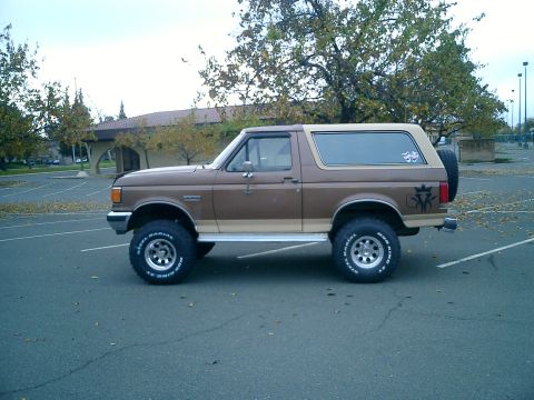 1990 Ford Bronco - This lifted Ford Bronco is parked in an empty lot with trees in the background.