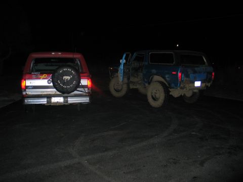 1995 Ford Bronco - Two full-size Ford Broncos parked on a dirt lot at night. At left, a red 1990s Bronco faces away showing a spare-tire cover reading FORD BR…