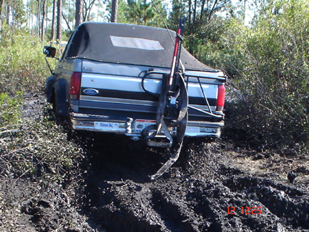 1992 Ford Bronco - Rear view of a blue and silver two-tone 1980s-90s Ford pickup with a black bed cap and a hi-lift jack strapped to the tailgate, buried in d…