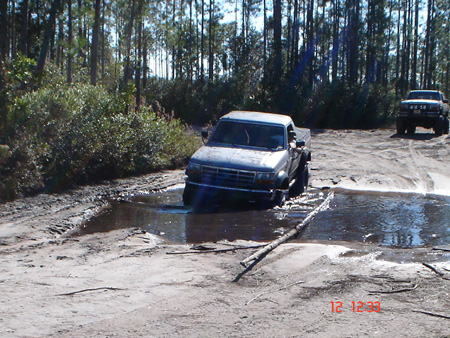 1992 Ford Bronco - A lifted 1992-1996 Ford truck with a mud-streaked body works through a puddle in a sandy rut on a pine-forest trail, with logs laid across…