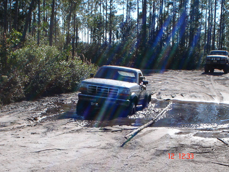 1992 Ford Bronco - A 1990s Ford pickup with a two-tone body splashes through a muddy puddle on a sandy pine-forest trail, water spraying from the rear tires.…