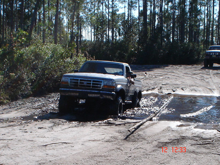1992 Ford Bronco - A lifted 1992-1996 Ford truck with a chrome grille and mud-caked lower body drops into a shallow water crossing on a sandy pine-forest trai…