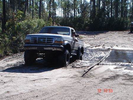 1992 Ford Bronco - Front three-quarter view of a lifted 1992-1996 Ford truck on mud tires, its lower body caked in dried mud, parked at the edge of a shallow…