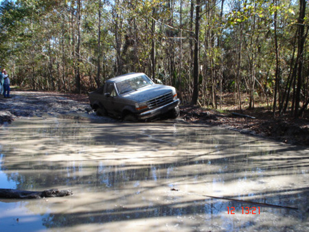 1992 Ford Bronco - A mud-covered 1990s Ford truck sits nose-down at the far edge of a wide, muddy water hole on a wooded dirt trail, with a bystander watching…