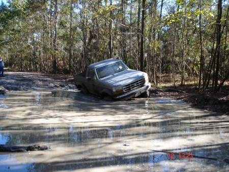 1992 Ford Bronco - A mud-caked 1990s Ford pickup with a regular cab and short bed sits tilted at the edge of a large water-filled mud hole on a wooded trail,…