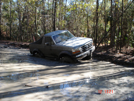 1992 Ford Bronco - A 1992-1996 Ford Bronco with its rear top removed sits axle-deep in a large muddy water hole on a wooded trail, body panels streaked with m…