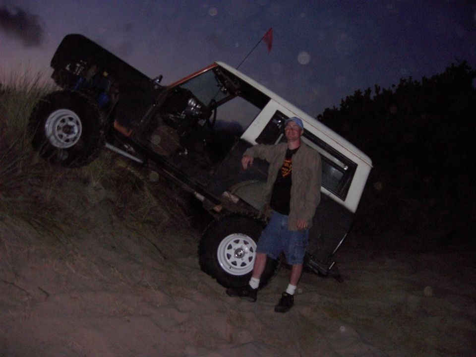 Man leaning on a doorless off-road Bronco parked on a sand dune at dusk
